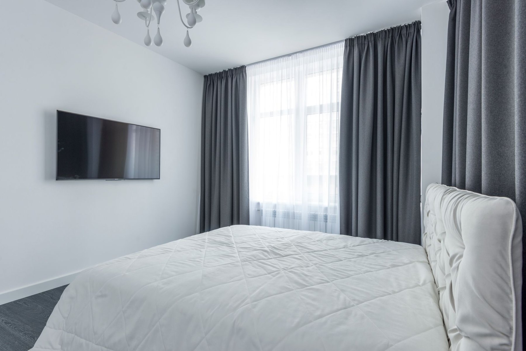 Bedroom with a white bed, dark gray curtains framing a sheer-covered window, and a wall-mounted television.