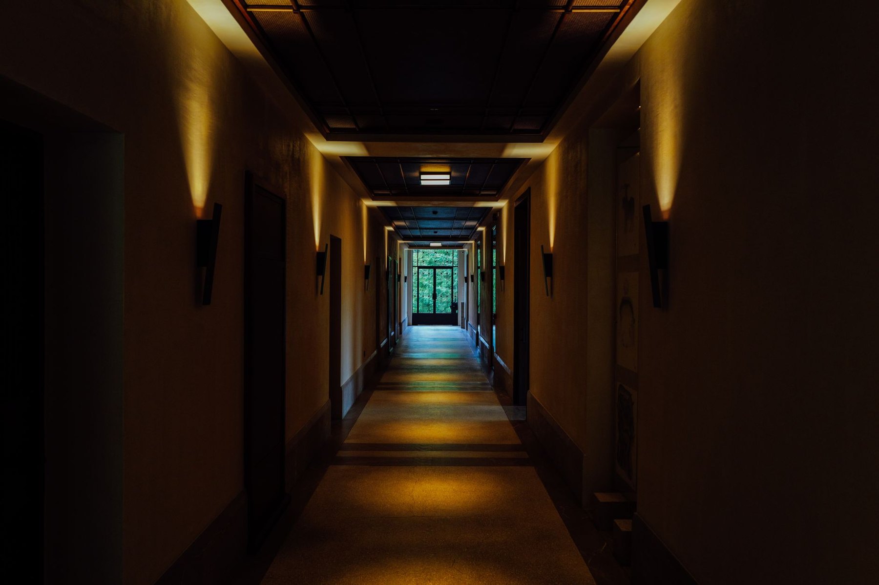 Dim corridor with warm wall lights illuminating the floor and doorways along both sides.