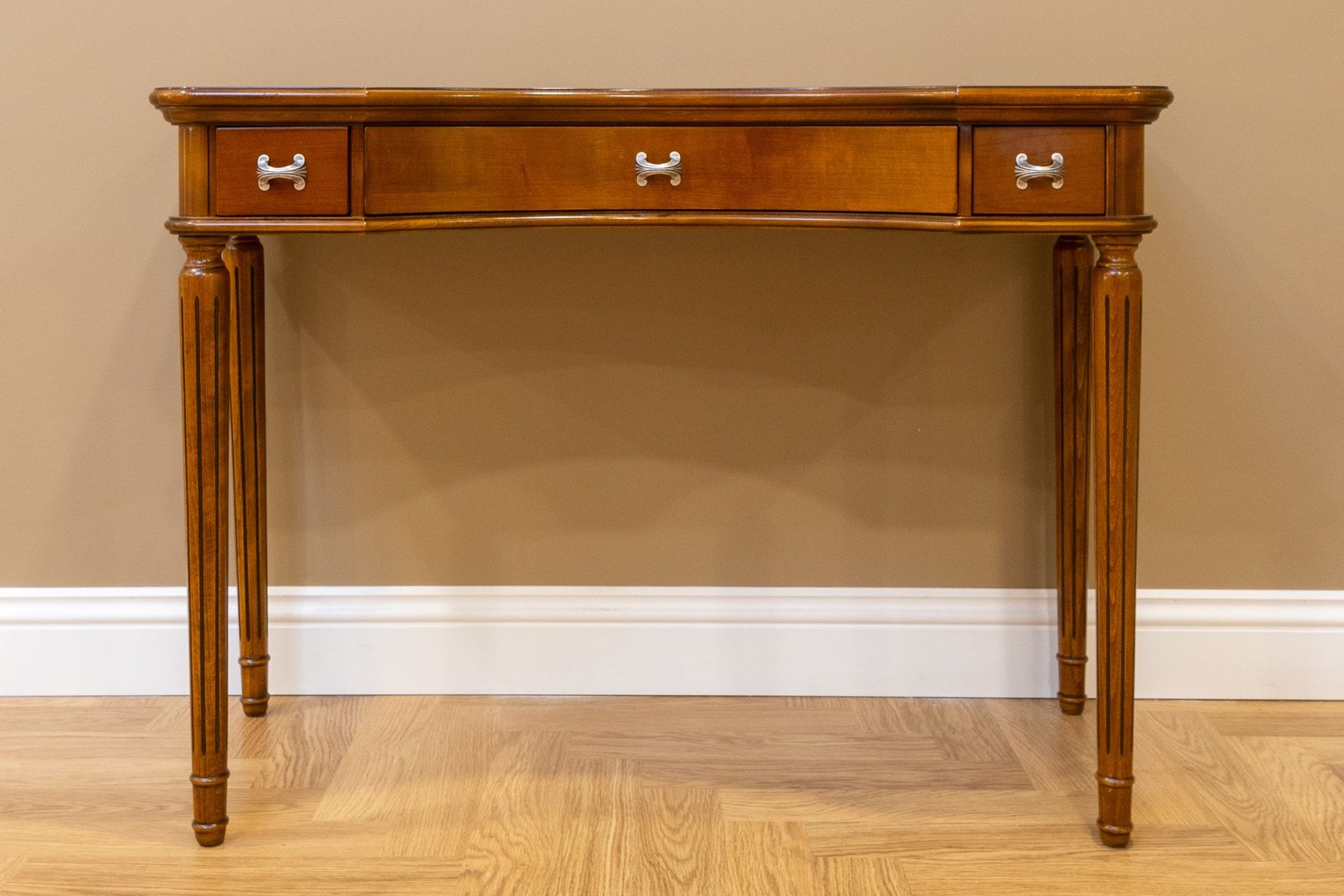 Wooden console table with three drawers, silver-colored handles, and four slender straight legs against a beige wall.
