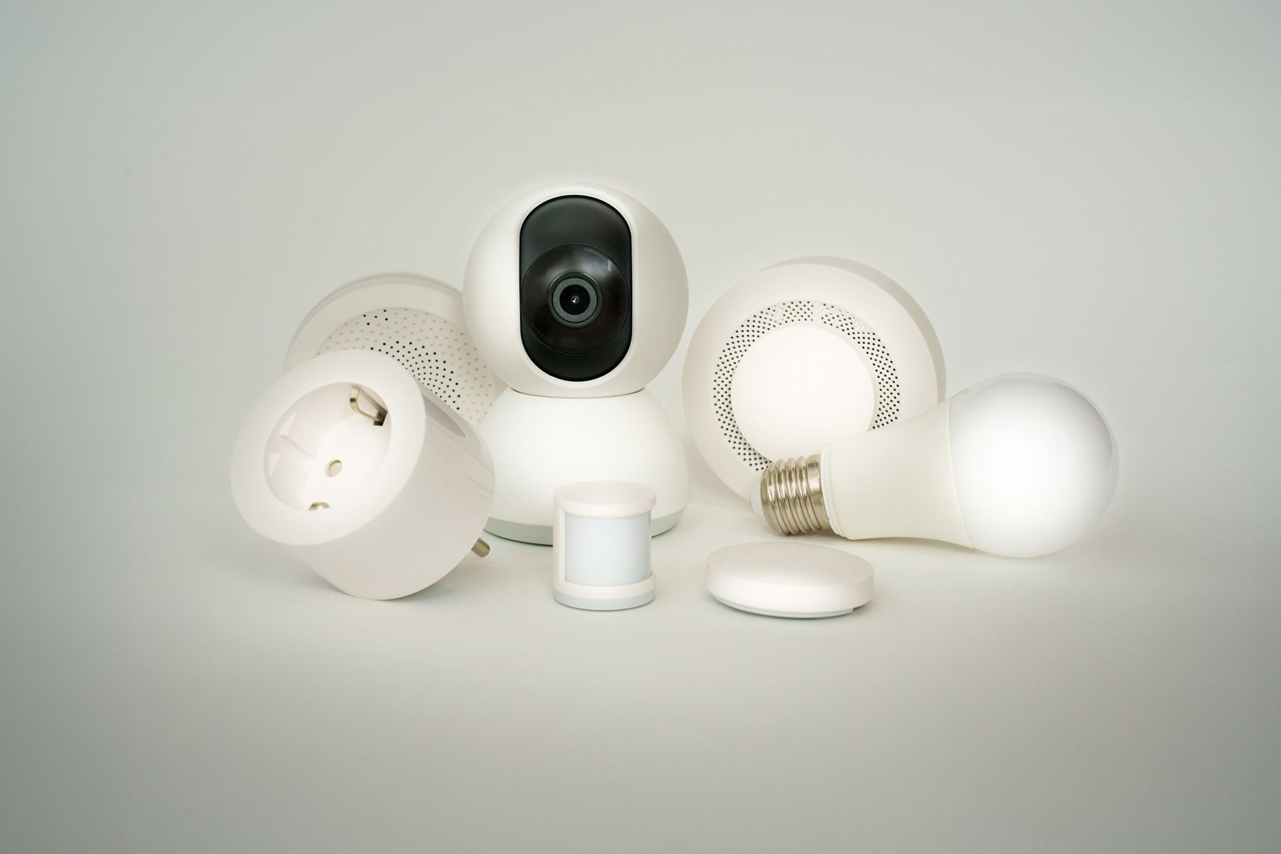 White smart home devices grouped together, including a camera, plug-in socket, light bulbs, and small sensors.